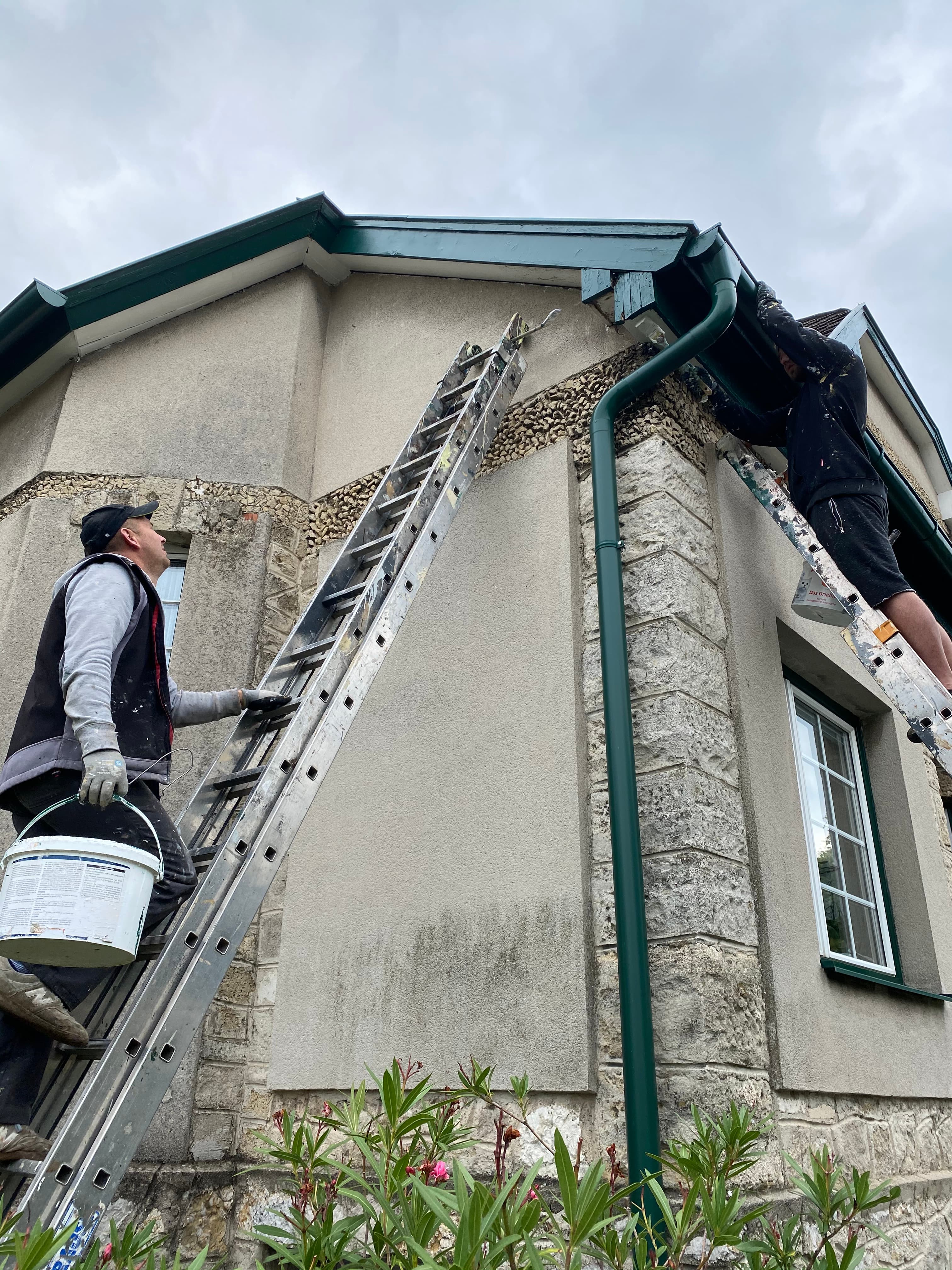 Zwei Männer auf Leitern führen Malerarbeiten an der Fassade und Dachrinne eines Hauses durch.