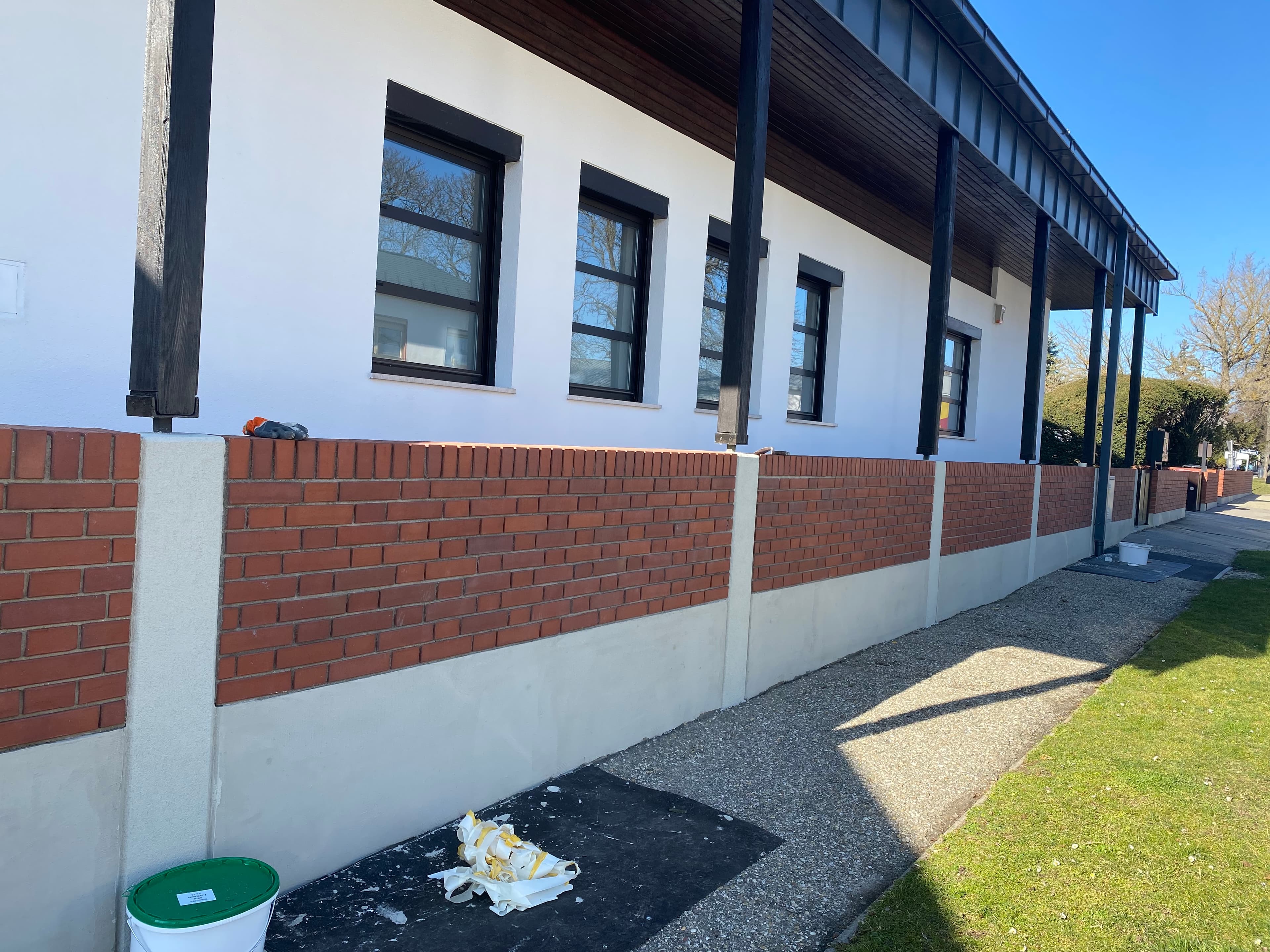 Moderne weiße Hausfassade mit schwarzen Fenstern hinter einer langen Ziegelmauer an einem sonnigen Tag.