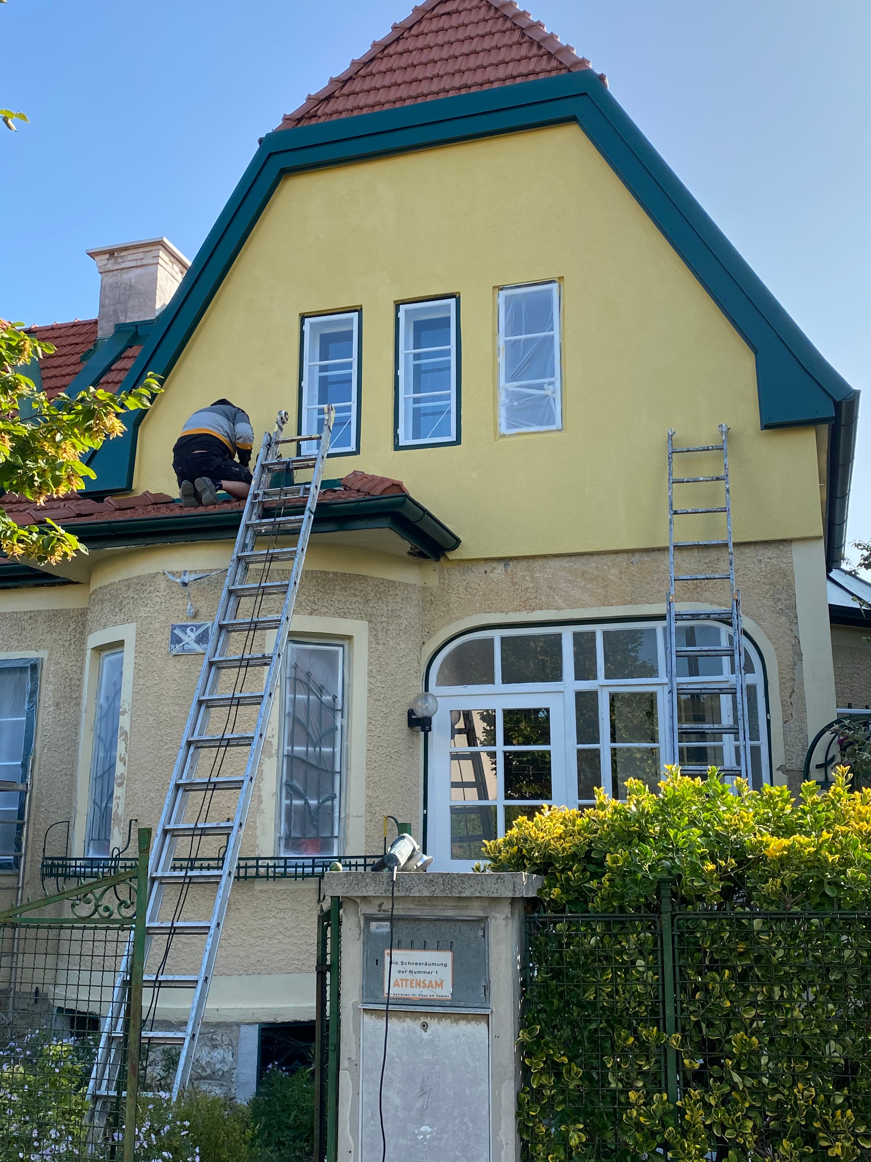 Handwerker auf dem Dach eines gelben Hauses mit grünen Akzenten und zwei langen silbernen Leitern.