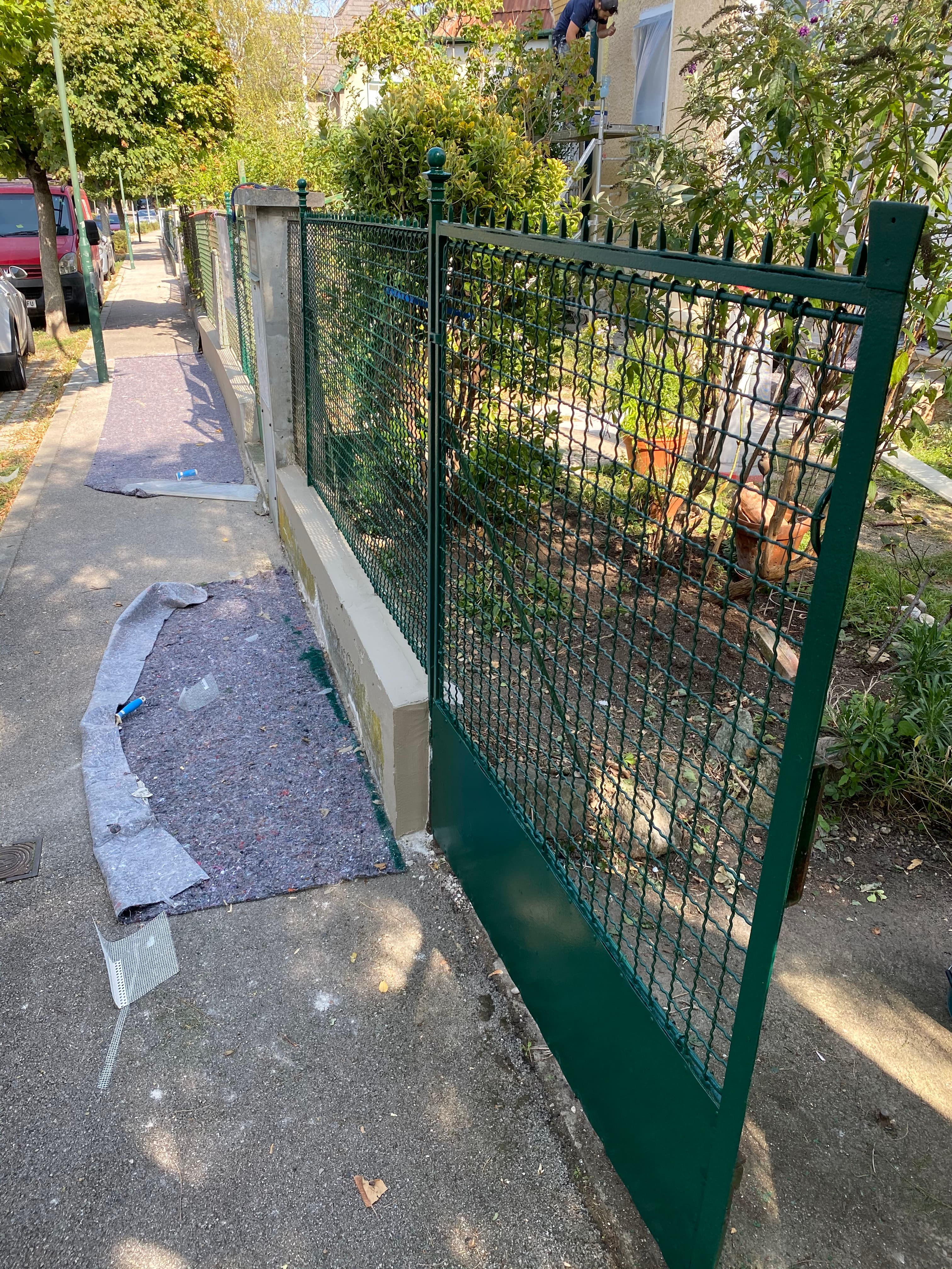 Offenes grünes Metalltor an einem Gehweg mit ausgelegtem grauen Malervlies vor einem grünen Garten.