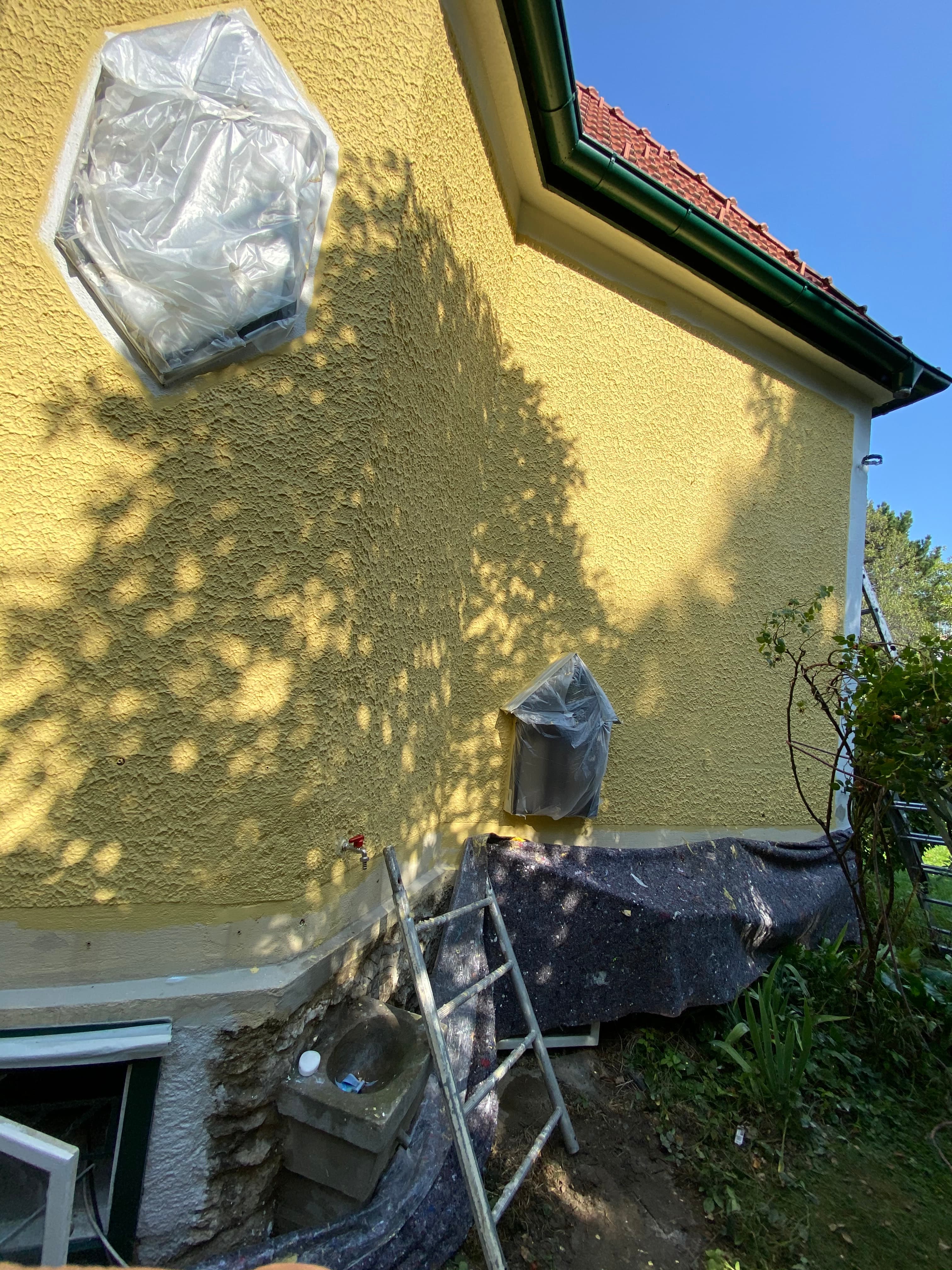 Gelbe Hauswand mit abgeklebten Fenstern, einer Leiter und Schutzplanen während Renovierungsarbeiten im Freien.