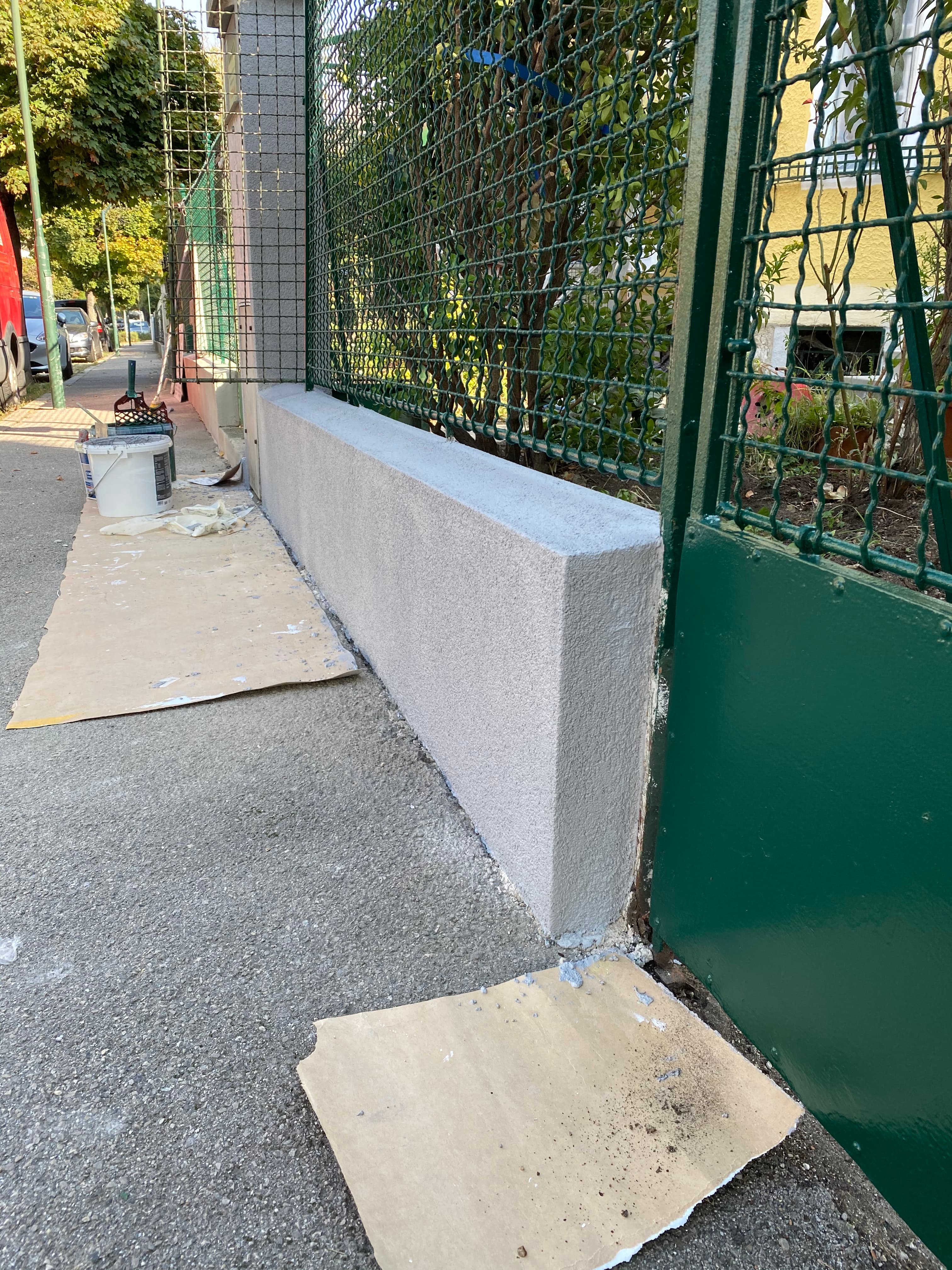 Frisch verputzter grauer Sockel eines grünen Metallzauns auf einem Gehweg mit schützender Pappe.
