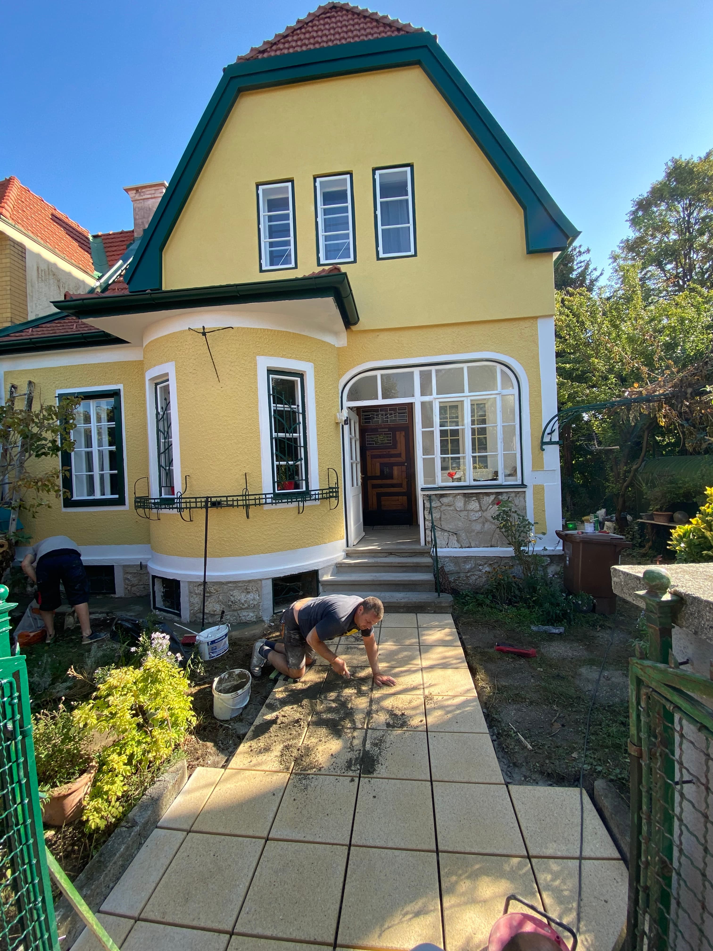 Ein Mann verlegt Steinplatten für einen Gehweg vor einem gelben Haus bei sonnigem Wetter.